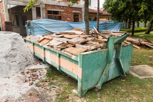 A large, rectangular industrial-style skip container made of metal with a weathered green finish, positioned on a grassy area next to a stone-paved pathway in a residential or semi-development setting. The skip is filled with a substantial amount of wooden debris, including planks, boards, and some smaller wooden fragments, appearing rough and untreated with a natural light brown to tan coloration. The skip's sides are reinforced with orange reflective stripes, and it has metal lifting points on the top edges, indicating it is ready for rubbish collection or clearance. In the background, there is a partially constructed or under-renovation building with exposed brickwork and scaffolding, along with blue tarpaulins covering parts of the construction site. A few trees with green foliage are visible, suggesting the setting is in a temperate climate. The environment is well-lit with natural daylight, highlighting the textures of the wood and the worn surface of the skip, aligning with rubbish removal services focusing on waste clearance in Battersea or nearby areas.
