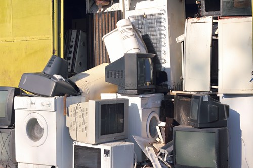A large collection of discarded household electronic appliances and small furniture items piled together outdoors, including washing machines, CRT monitors, microwave ovens, and a toaster, some placed atop others to form a dense heap on a cement surface. The appliances are made of metal, plastic, and glass, with visible textures such as smooth, glossy, and matte finishes, predominantly in white, black, grey, and beige tones. The background features a weathered yellow metal structure and a dark, partially open storage unit or shed, indicating an outdoor clearance or junk collection area near Battersea, where Battersea House Clearance operates. The sunlight casts natural shadows, highlighting the cluttered arrangement of the rubbish. This image represents typical household waste collected for rubbish removal services, illustrating the type of items handled during house clearance operations in the Battersea region.