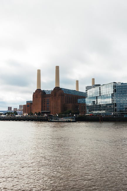 A large industrial building with a red brick facade featuring vertical architectural details and multiple rectangular windows, situated along a riverbank. The building has three tall, cylindrical chim