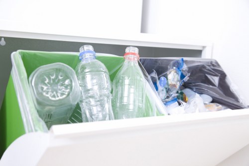Inside a white rubbish disposal unit, there are three visible compartments containing different waste items. The front compartment holds two clear plastic bottles, one with a blue cap and the other with a red cap, positioned horizontally and partially overlapping. Behind the bottles, a green plastic container is visible, possibly serving as a divider within the unit. To the right, a black plastic bag filled with discarded waste, including some small blue plastic items and other miscellaneous rubbish, is seen leaning against the side of the container. The background shows part of a light-colored interior space, suggesting an indoor setting within a residential property. The waste appears to consist of recyclable plastic bottles and general waste, typical of rubbish removal services such as Battersea House Clearance, operating around the Battersea and Wandsworth areas. The overall scene is well-lit, with natural or artificial light highlighting the textures and colors of the waste materials, emphasizing the importance of proper waste segregation during rubbish clearance activities.