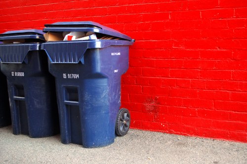Two large dark blue plastic wheelie bins with black lids are positioned on a concrete pavement in front of a vibrant red brick wall. The bins are designed for rubbish collection, with textured surfaces and visible wheels at the rear for mobility. One bin is slightly taller and wider than the other, and they are placed closely together, with the larger bin partially overlapping the smaller one. The lids are closed, and the bins are situated outdoors, possibly on a residential driveway or communal area in Battersea, London, given the postcode inference. The rough concrete ground beneath shows subtle speckling and texture, contrasting with the smooth, painted brick wall behind. The bright red bricks are evenly laid and create a striking background, emphasizing the utilitarian presence of the bins, which are part of a rubbish removal service often provided by companies like Battersea House Clearance. The natural lighting suggests a clear, daytime setting, with no additional equipment or action visible in the scene.