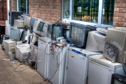 A large collection of discarded electronic waste, including various old computer monitors, desktop towers, and other obsolete electronic components, is lined up outside against the exterior brick wall of a building. The monitors and towers are of different sizes and styles, with some featuring curved screens and others flat panels, in shades of grey, beige, and black. Many of the units show visible cables and wires, with some power supplies and internal components partially exposed. The electronic waste is arranged on the ground, which appears to be concrete, next to a window with a black frame and reflective glass pane. The scene is illuminated by natural daylight, creating subtle shadows across the objects, emphasizing the textures of the aging plastic casings and metal parts. This arrangement of electronic waste reflects the type of clutter often managed by Battersea House Clearance during rubbish removal services, particularly in urban areas like Battersea, London postcode SW11, where disposal of obsolete electronics is a common task.